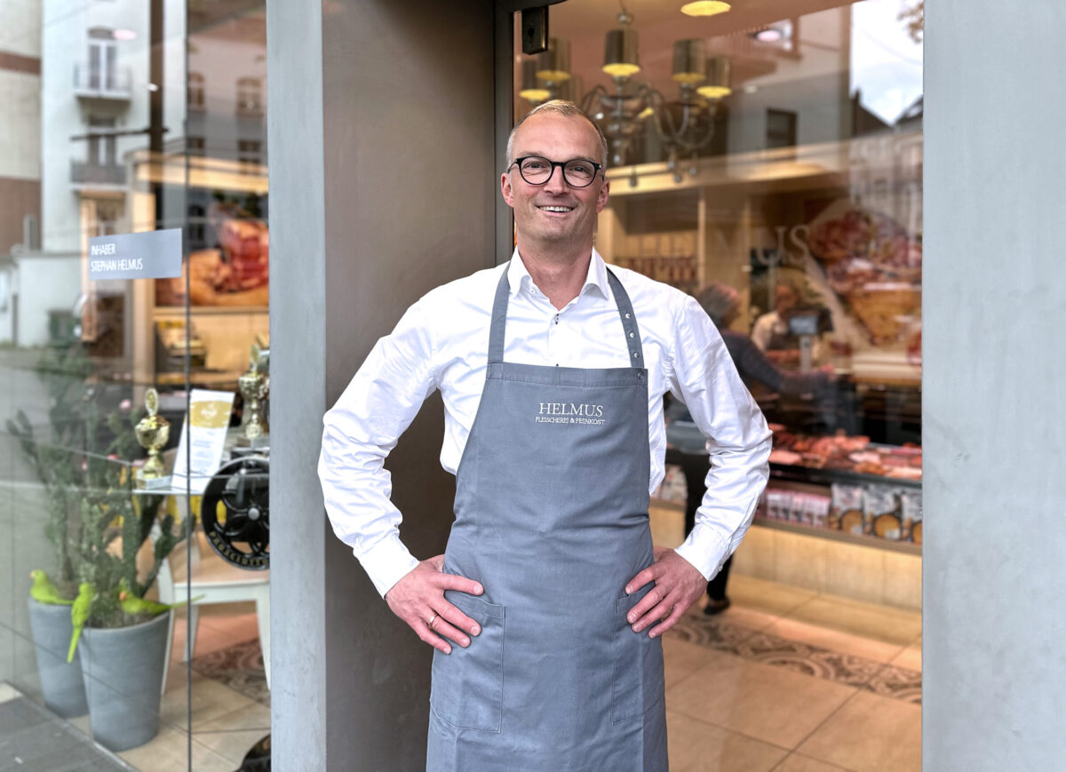 Metzgermeister Stephan Helmus lächelt vor seiner Metzgerei in die Kamera. 