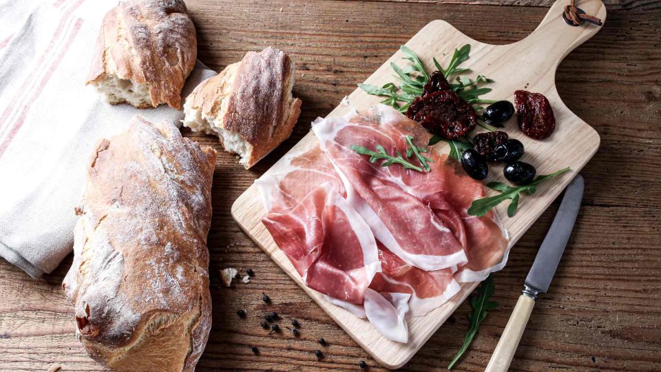 Parmaschinken auf einem Holzbrett, umgeben von Oliven und frischem Baguette. 